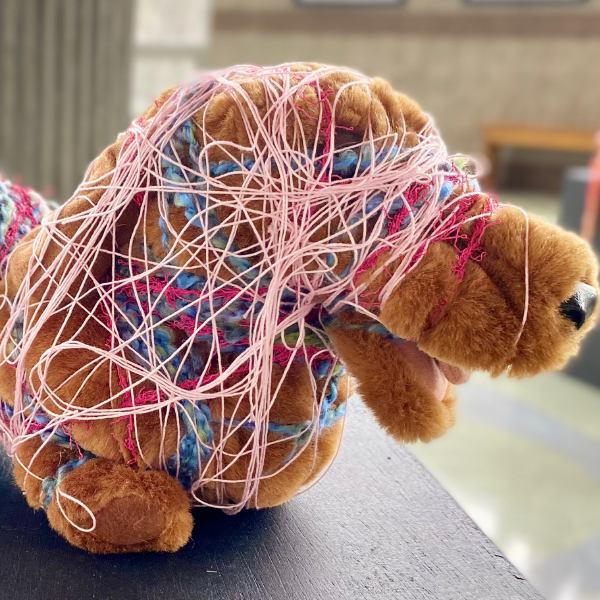 A small brown stuffed dog is wrapped in various materials. The head is wrapped in an abstract pattern with a thin, light pink string. The body of the dog is wrapped with various materials including a straw hat and multi-colored strings of yarn