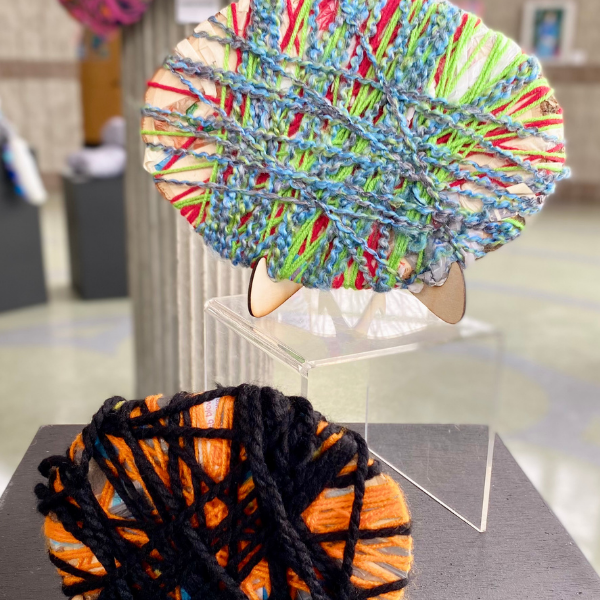 Two oval disks, approximately 10 and 8 inches in diameter. The larger disk is covered in an overlapping pattern of plastic bags strands and blue, red, and green yarn. The smaller disk is covered in an overlapping pattern of plastic bags and black and orange yarn.