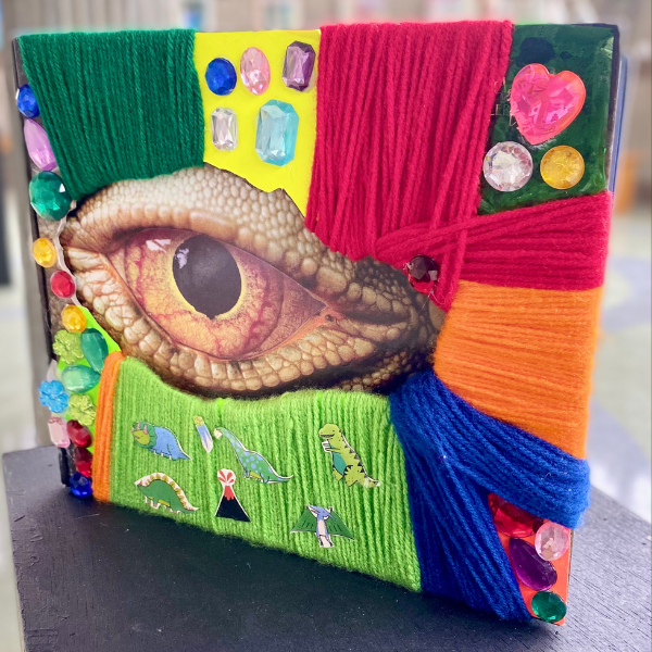 A hardback book that features the cutout shape of a reptilian looking eye in shades of orange and beige. The outside cover is lined in neat color blocked rows of yarn in shades of blue, green, red and orange. Interspersed between the yarn are clusters of gemstones in various colors and small dinosaur stickers. The back of the book is also wrapped in neat rows of red, purple and green yarn. Woven into the yarn are six plastic miniature dinosaur toys in green and orange.
