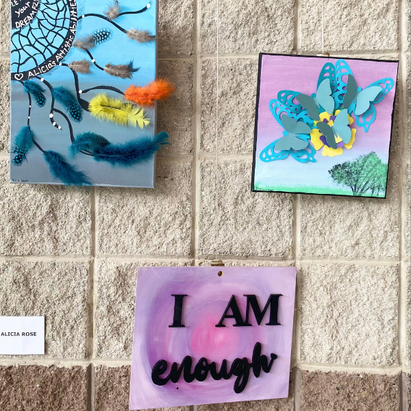 A trio of small paintings. One features a black dream catcher web in the upper left corner. The background is an ombre of blue and gray. In white lettering within the dream catcher it reads “Let you dreams fly” and “Alicia’s Artistic Abilities”. There are strands of colorful three dimensional blue, yellow, and orange feathers streaming from the catcher. The second painting features a collection of overlapping three dimensional butterflies in shades of blue, yellow, and gray. The backdrop of the painting is blue and purple with the silhouette of a patch of trees in the left corner. The third painting is a swirling pink and purple background with raised black lettering that reads “I AM enough.”