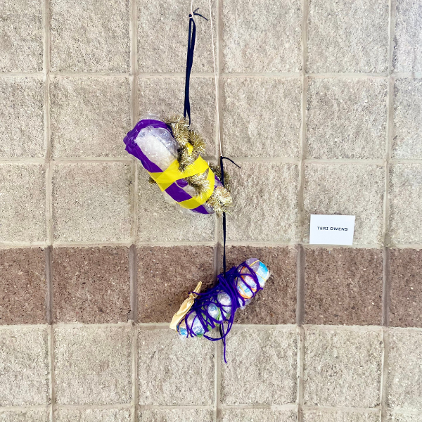A skinny dowel rod is suspended in the air. Hanging from the rod is a string and black laces with two shoe-shaped objects hanging from it. The objects are covered in multiple overlapping layers of material: bubble wrap, yellow plastic tape, gold garland, purple tape, and purple yarn.